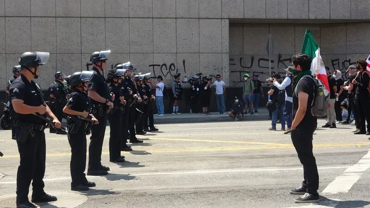洛杉磯數百人群眾聚集在聯邦政府大樓前，抗議移民暨海關執法局（ICE）突襲掃蕩非法移民，一名示威者手拿墨西哥國旗，在一排警察面前站立表示抗議。 中央社