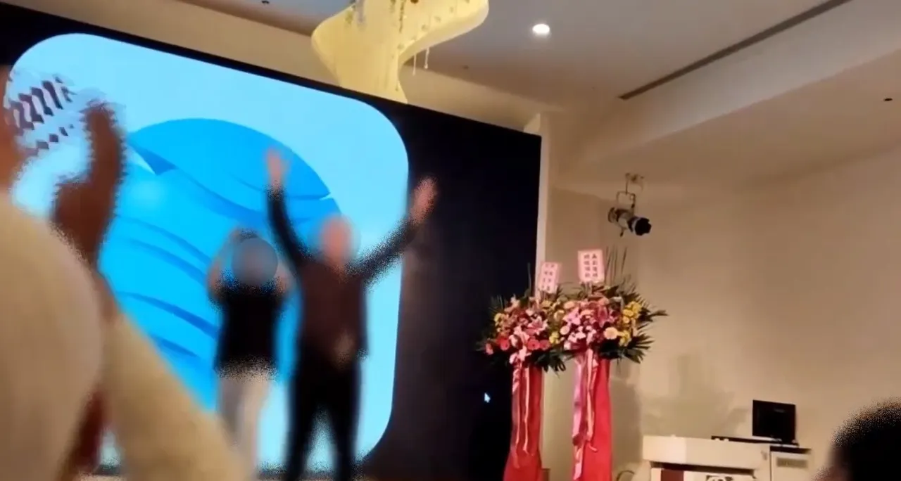 股票老师丁教授一路从台下走上台,与群众们击掌欢呼,没想到一切都是诈团演出来的。翻摄画面