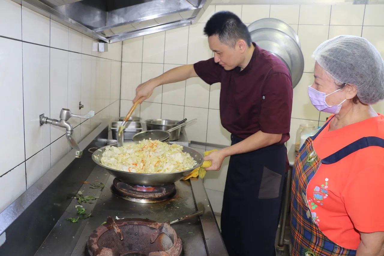 五星级饭店主厨的陈佳玮专程到新庄牡丹心社区发展协会照顾关怀据点为长辈义煮,志工在一旁讨教炒菜好吃秘诀。社会局提供