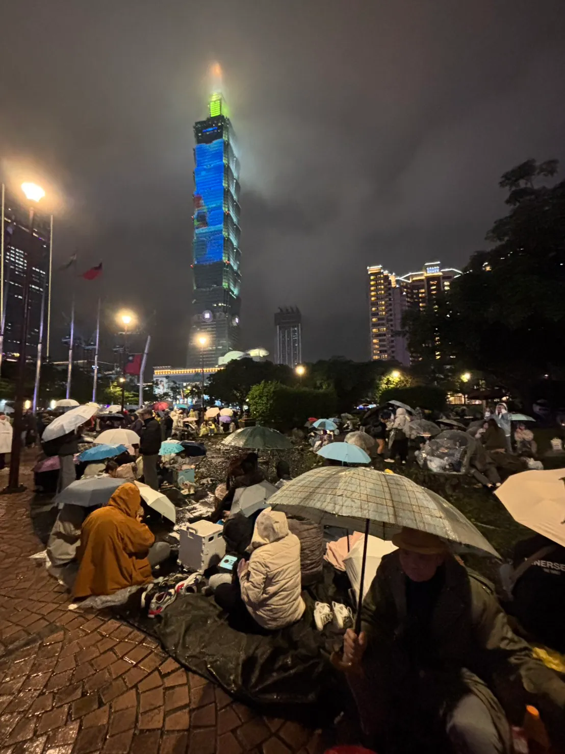 民众聚集在府前广场的公园内，打伞席地而坐等待101烟火。江孟谦摄