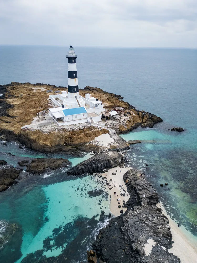 黑白相間的塔身矗立在黑褐色的玄武岩礁上，與碧綠清澈的海水形成強烈對比。田欣雲攝